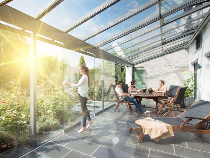 Terrassendach mit Glas-Faltwand, die gerade von einer Frau geöffnet wird. Die Sonne scheint und im Hintergrund sitzen zwei Leute an einem Tisch.