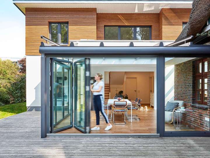 Wintergarten vor einen modernen Wohnhaus. Die Glas-Faltwand wird gerade von einer Frau geöffnet.